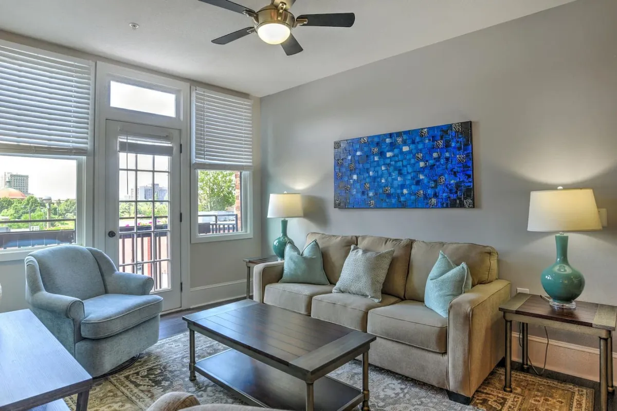 The image showcases a stylish, inviting living room with a comfortable sofa, tasteful artwork, ambient lamps, and large windows that fill the space with natural light. The setting evokes warmth and relaxation, perfect for gathering with friends or enjoying quiet moments as the sun sets over Asheville.