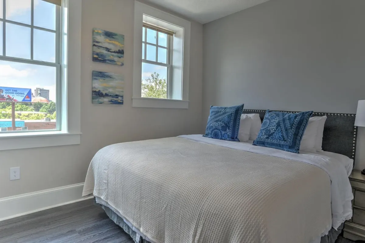 A cozy bedroom with decorative pillows, striking wall art, and expansive windows that flood the space with natural light. The setting exudes tranquility, offering a perfect spot for relaxation, reading, or simply enjoying the peaceful ambiance.