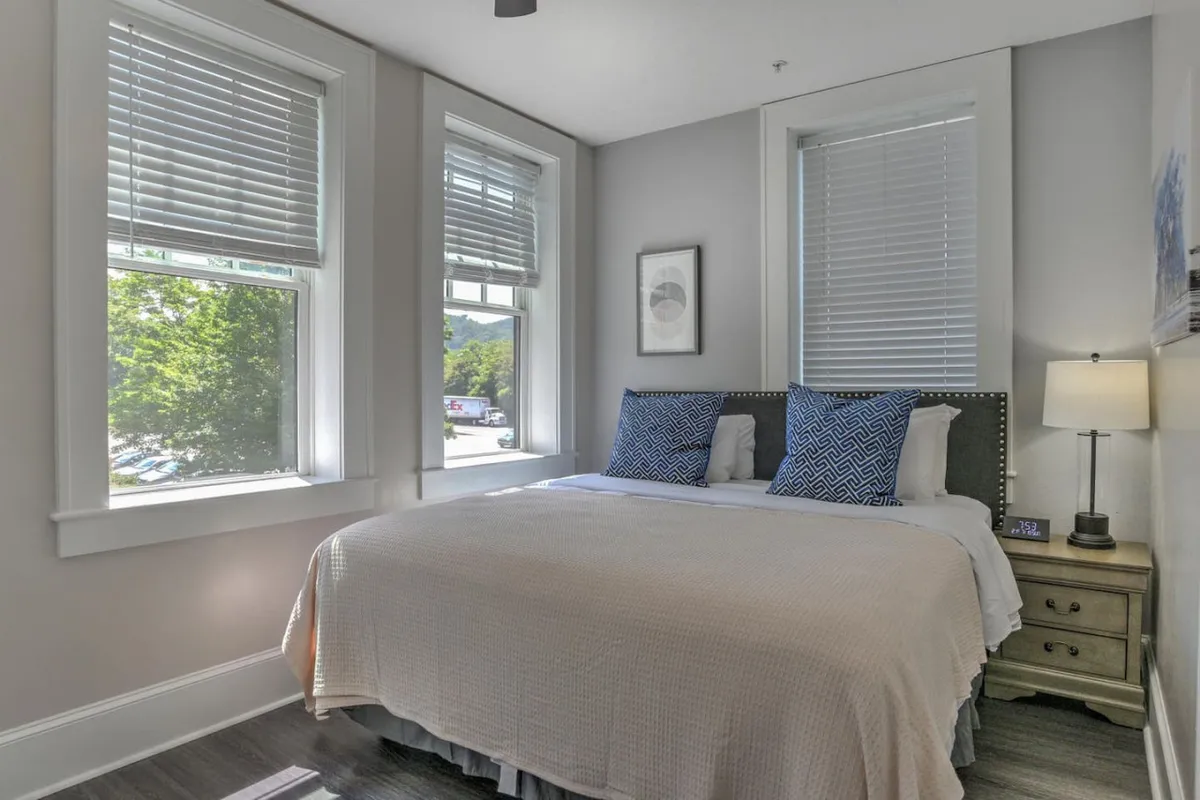 A serene bedroom featuring a comfortable bed adorned with decorative pillows, flanked by a nightstand and lamp. Large windows allow gentle natural light to stream in, creating an inviting and restful atmosphere perfect for unwinding or leisurely mornings.
