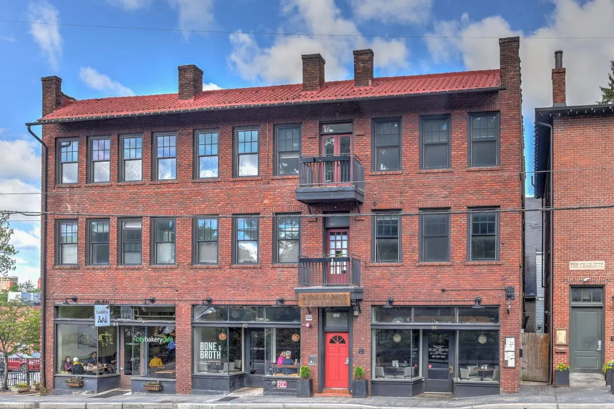 An exterior shot highlighting a red door entrance, large windows, a brick facade, and a balcony. The scene feels warm and welcoming, hinting at the boutique stores and neighborhood cafes just steps away, and evokes the pleasure of sunny afternoons enjoyed outdoors.