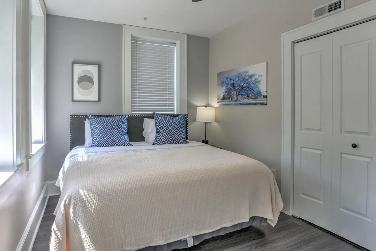 A serene bedroom scene featuring a comfortable bed with blue accent pillows, complemented by tasteful artwork and a softly glowing lamp. The space is filled with gentle natural light, creating a calm and welcoming atmosphere for relaxation or quiet work.